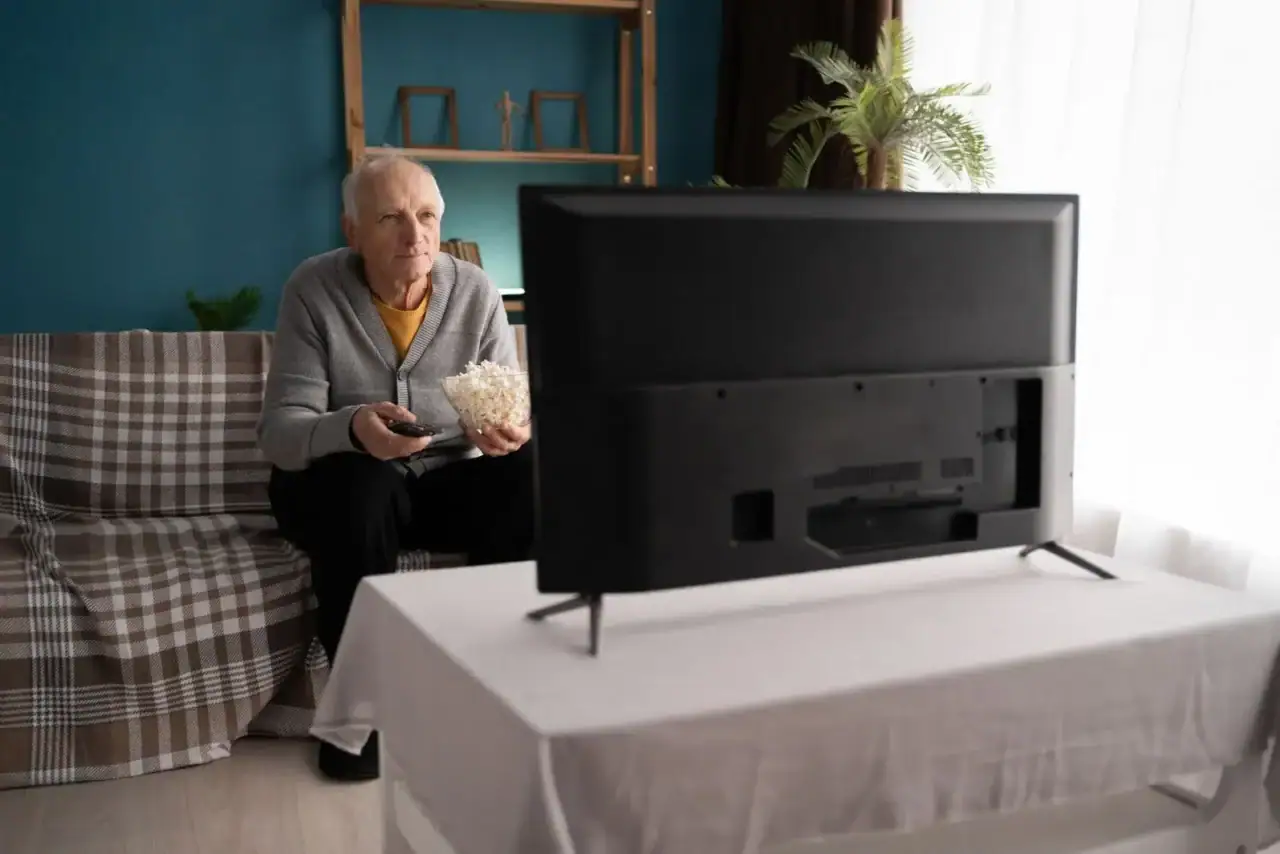 Older man on a plaid sofa watching TV while holding a bowl of popcorn and a remote.