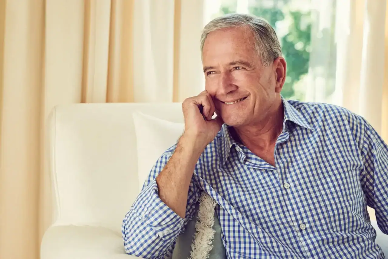 Older man smiling while talking on a phone, seated on a light sofa in a bright living room with cream curtains behind him.