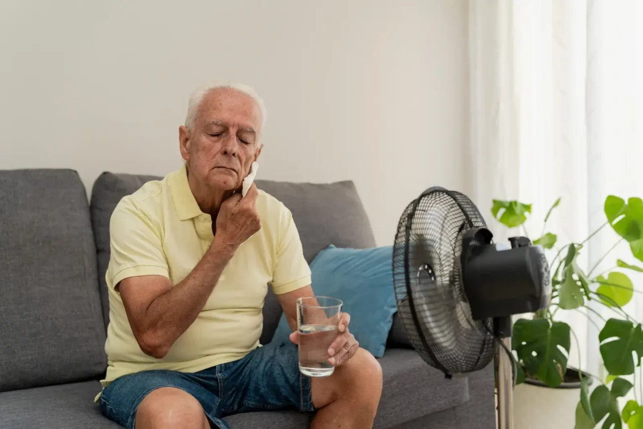 Senior se během horkého dne doma ochlazuje ventilátorem a doplňuje tekutiny.